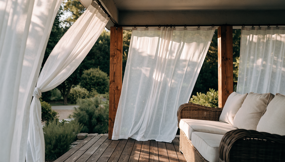 White outdoor sheer curtains moving in the breeze on a covered timber deck with a wicker sofa