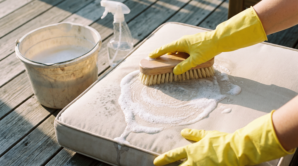 Hands in rubber gloves scrubbing mildew spots off a light outdoor cushion with a soft brush on a sunny patio