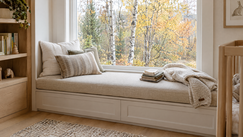 Cream window seat cushion with fabric swatches and a knit blanket beside a window with autumn birch trees