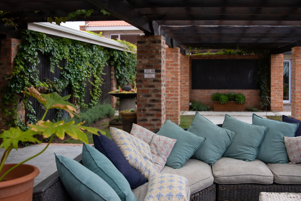 Wicker sectional with teal and patterned outdoor cushions under a pergola beside a brick wall